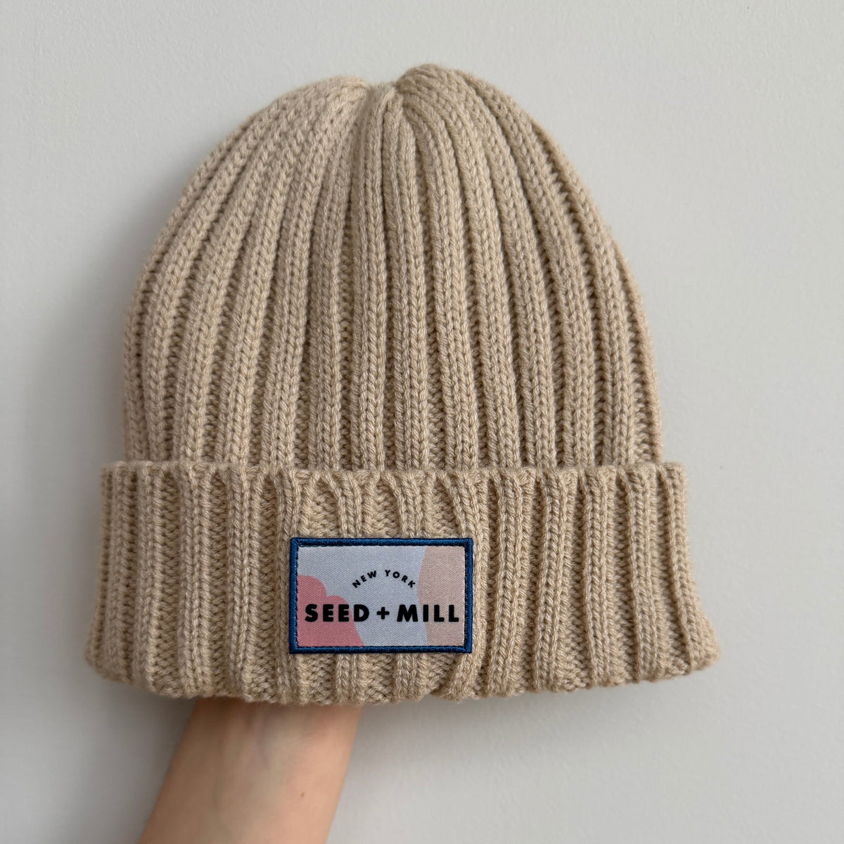 Beige knit beanie with a Seed + Mill patch on a light gray background
