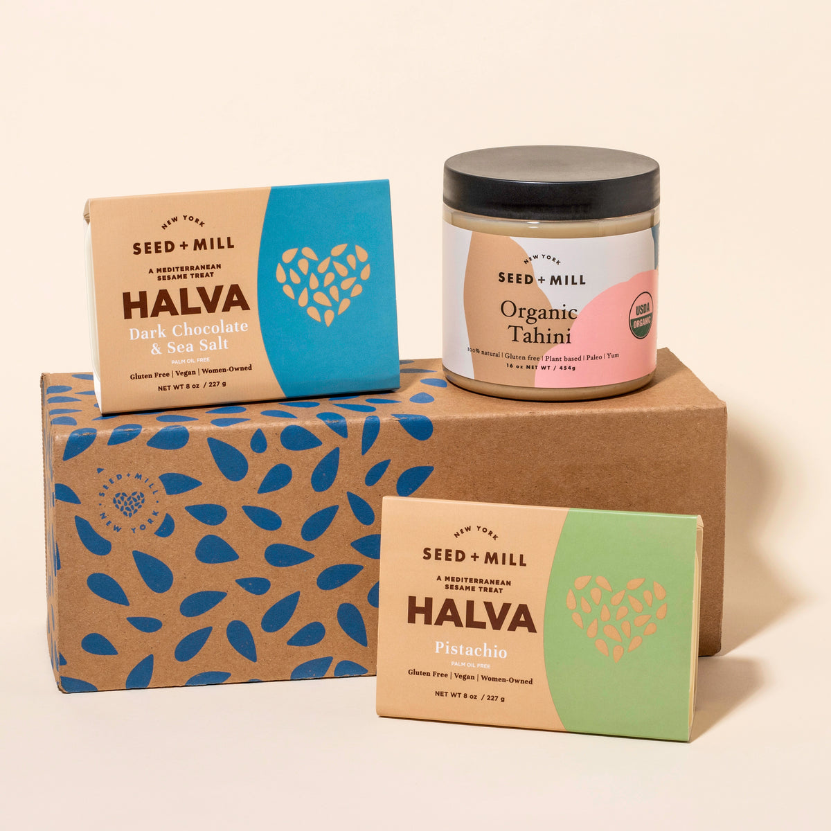 Assorted Seed + Mill products including a box of Sark Chocolate & Sea Salt halva, a box of pistachio halva, and a jar of organic tahini on a beige background.