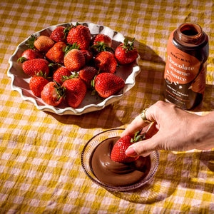 A plat of strawberries, a squeeze bottle of chocolate sesame sauce, and a hand dipping a strawberry into a bowl of chocolate sesame sauce