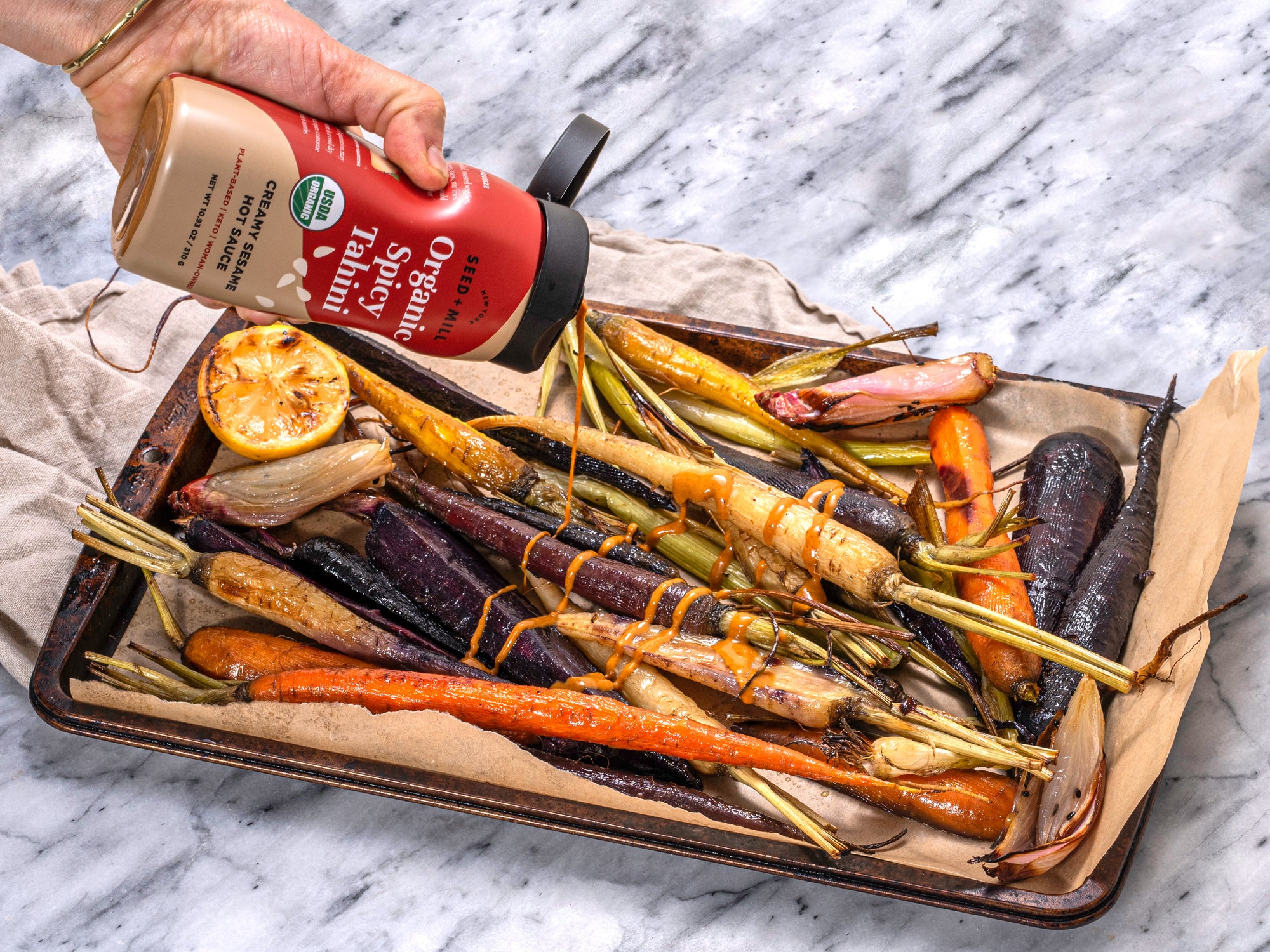 Roasted vegetables on a baking tray with a bottle of Seed + Mill Organic Spicy Tahini being squeezed over them.