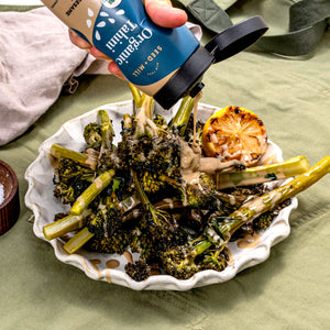 Person pouring Seed + Mill Organic Tahini over roasted vegetables on a plate with a green background