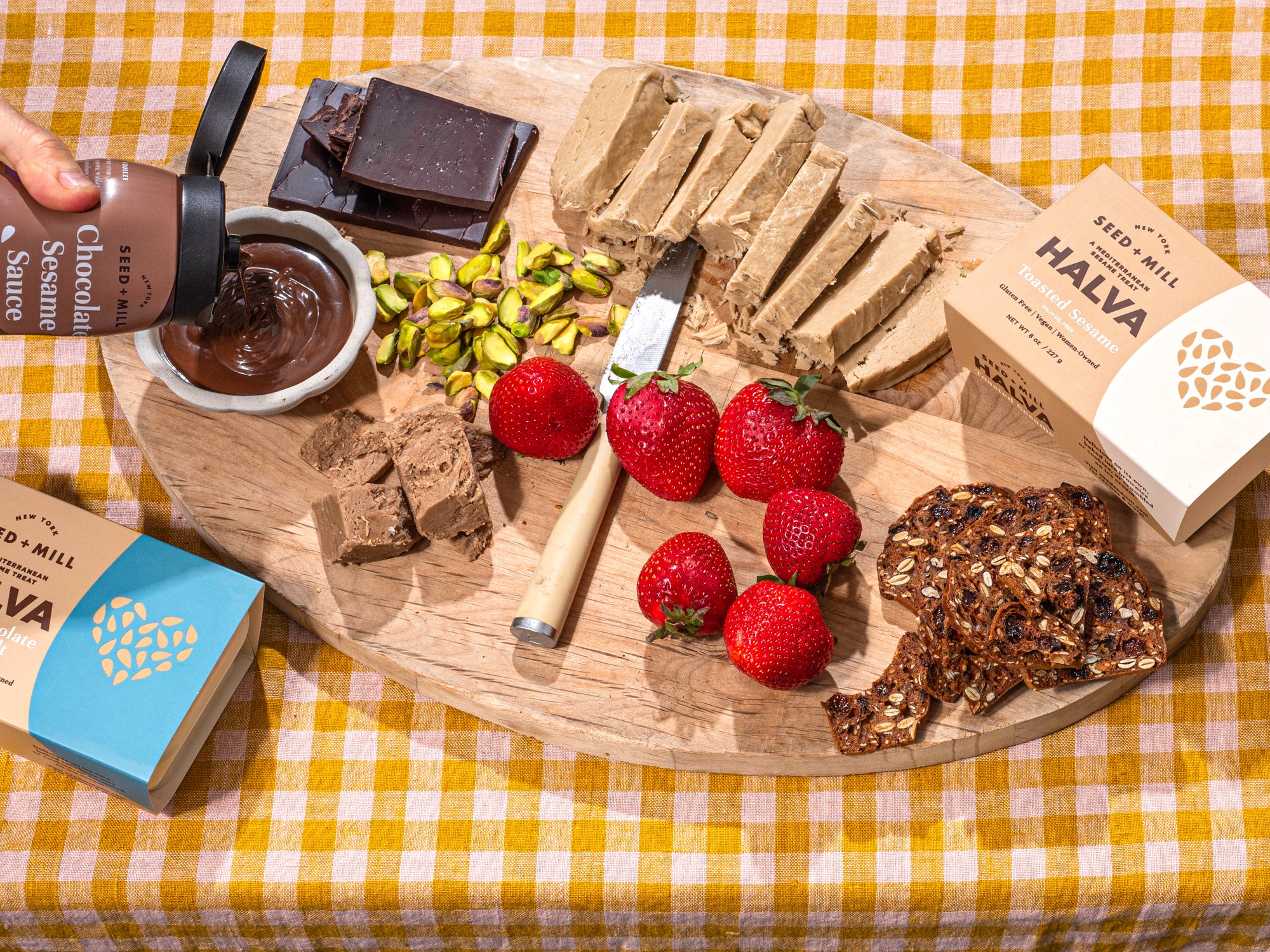 Wooden board with assorted foods including Seed + Mill Halva, Seed + Mill Chocolate Sesame Sauce, strawberries, chocolate, pistachios, and crackers. 