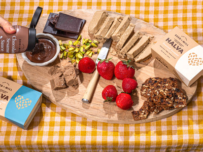 Wooden board with assorted foods including Seed + Mill Halva, Seed + Mill Chocolate Sesame Sauce, strawberries, chocolate, pistachios, and crackers. 
