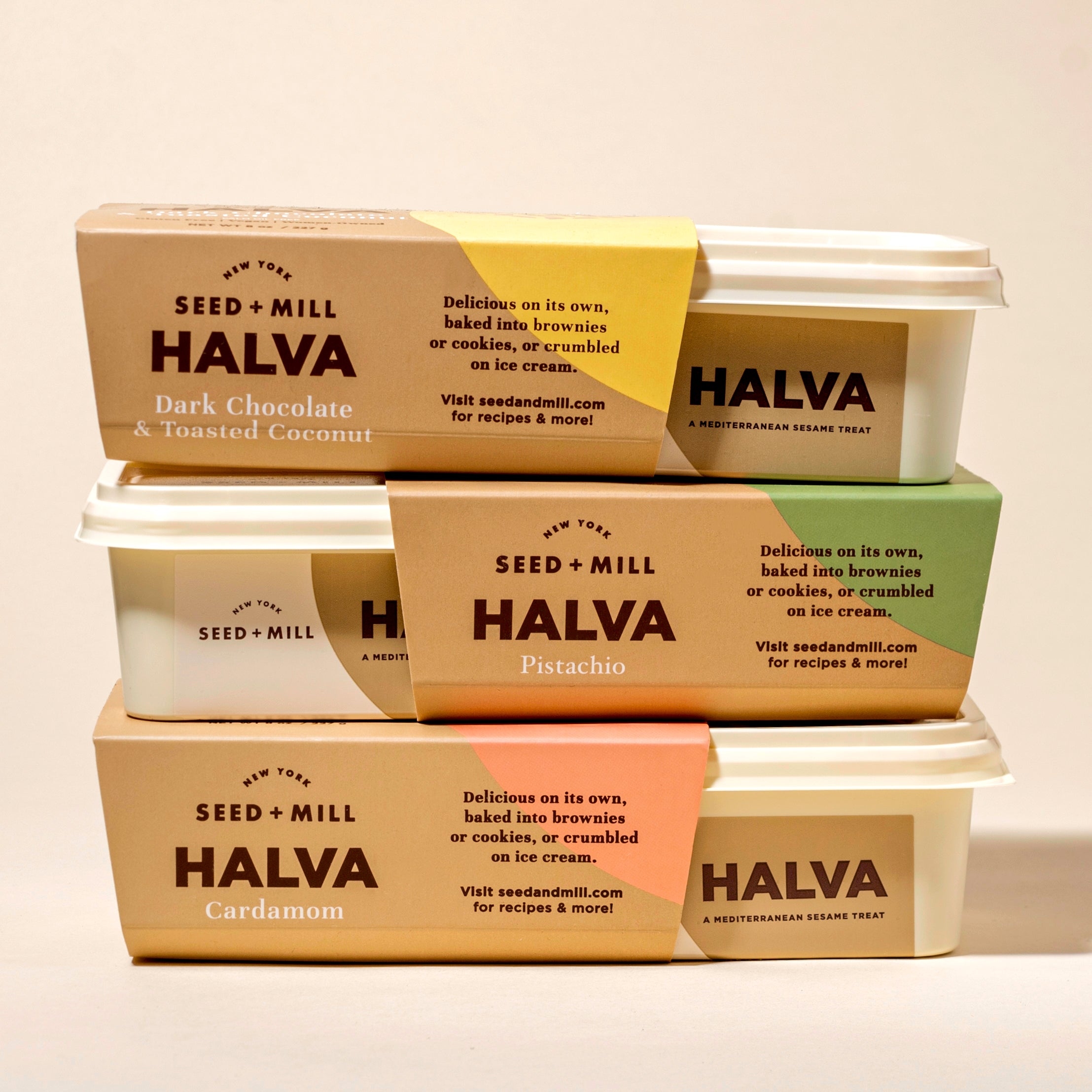 Stack of Seed + Mill Halva containers with various flavors on a beige background