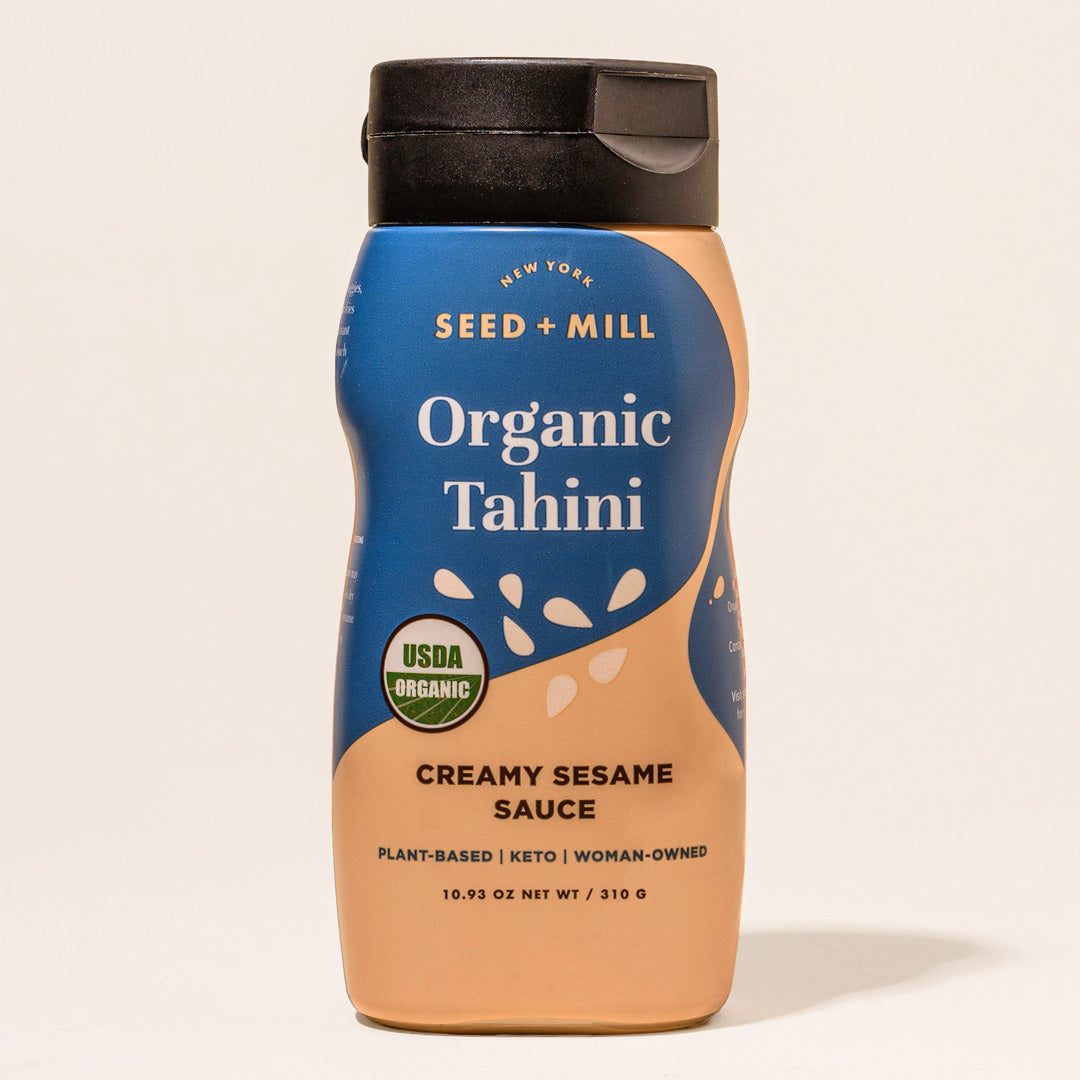 A bottle of Seed + Mill Organic Tahini with a blue label, displaying the product name and the description 'Creamy Sesame Sauce'. The bottle is upright and has a black cap.