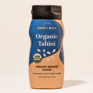 A bottle of Seed + Mill Organic Tahini with a blue label, displaying the product name and the description 'Creamy Sesame Sauce'. The bottle is upright and has a black cap.