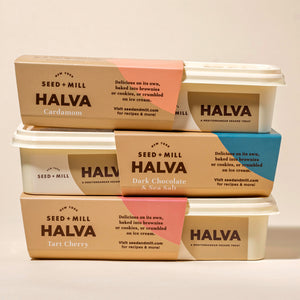 Stack of Seed + Mill Halva containers with various flavors on a light background