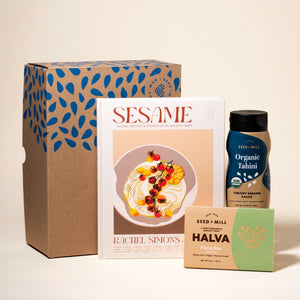 Collection of Seed + Mill products including a box of pistachio halva, Sesame (a cookbook), and a squeeze bottle of organic tahini on a beige background