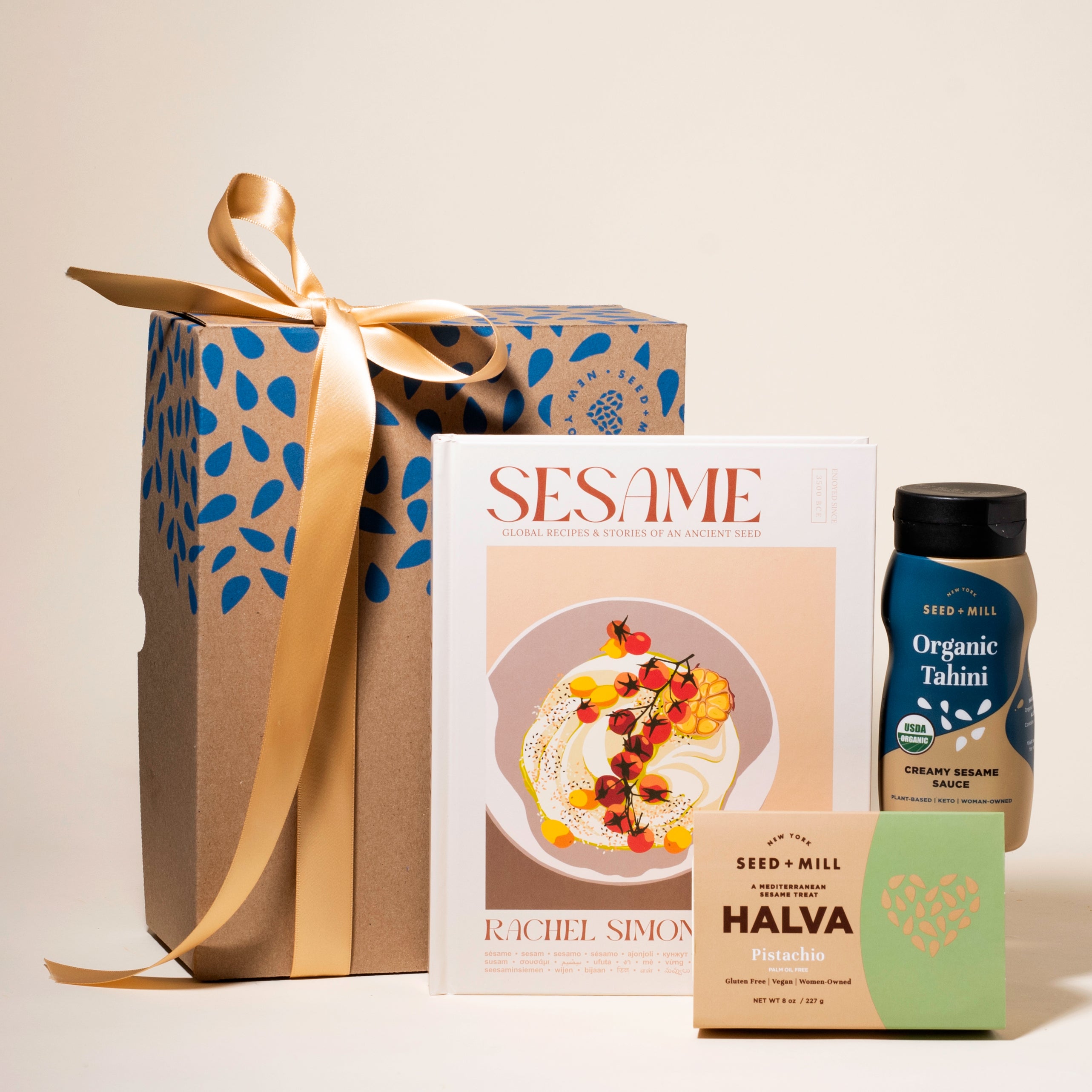 Gift box of Seed + Mill products including a box of pistachio halva, Sesame (a cookbook), and a squeeze bottle of organic tahini on a beige background