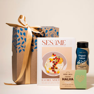 Gift box of Seed + Mill products including a box of pistachio halva, Sesame (a cookbook), and a squeeze bottle of organic tahini on a beige background