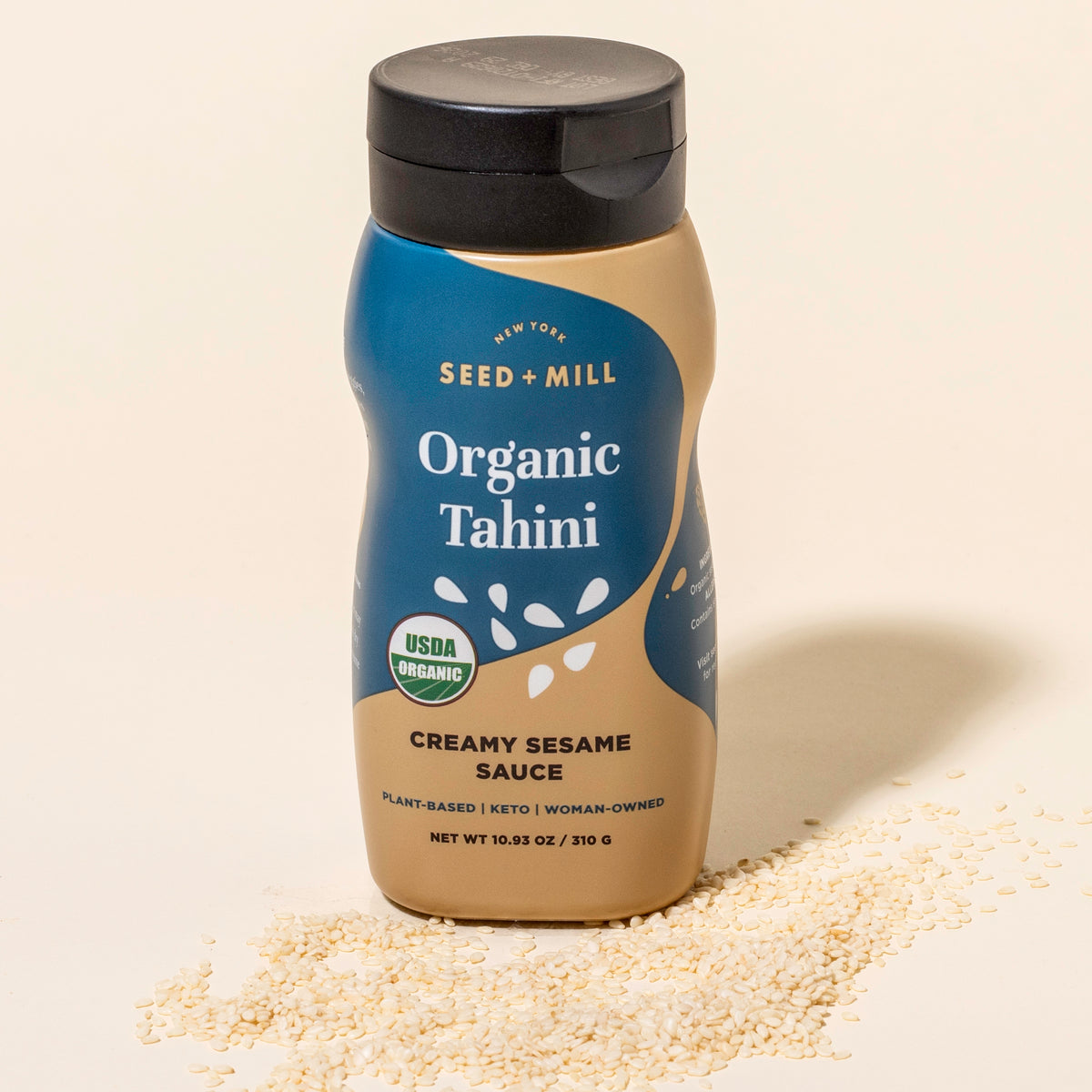 Bottle of Seed + Mill Organic Tahini on a beige background with sesame seeds around.