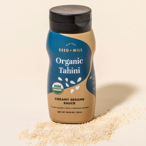 Bottle of Seed + Mill Organic Tahini on a beige background with sesame seeds around.