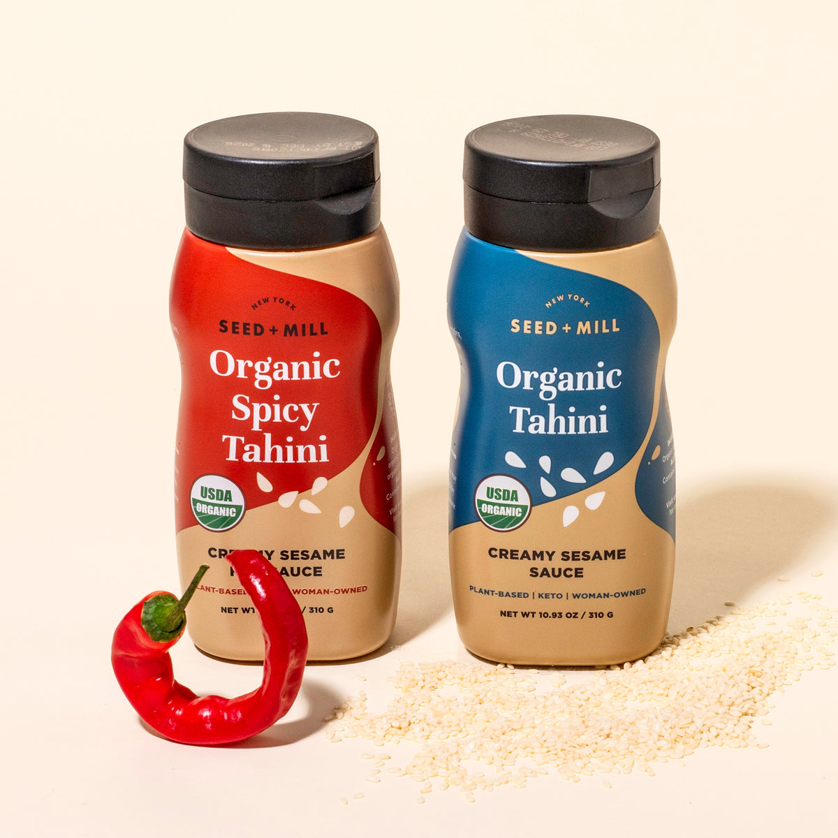 Two bottles of Seed + Mill Organic Spicy Tahini and Organic Tahini with a red chili pepper on a beige background.