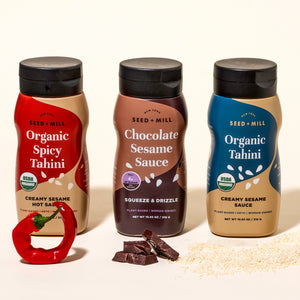 Three bottles of Seed + Mill Tahini, Organic Spocy Tahini, Chocolate Sesame Sauce, and Organic Tahini, with a red pepper and chocolate pieces on a beige background