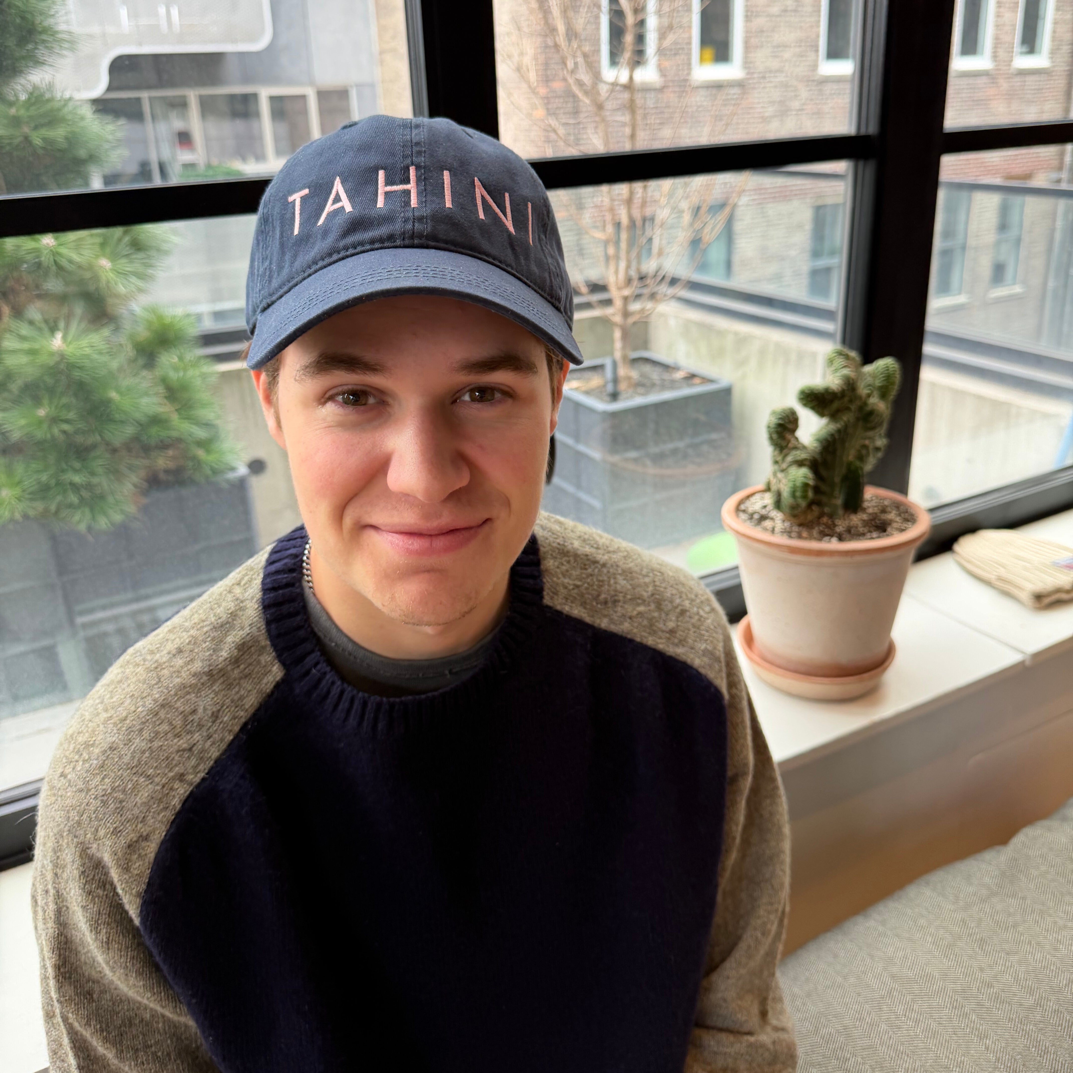 Person wearing a cap with 'Tahini' text, sitting indoors near a window with a cactus plant.