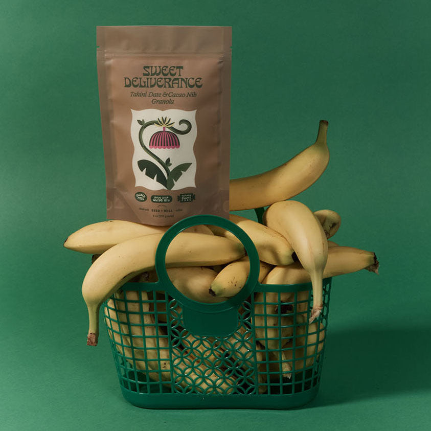 Sweet Deliverance Tahini Date & Cacao Nib granola package on top of a basket of bananas with a green background