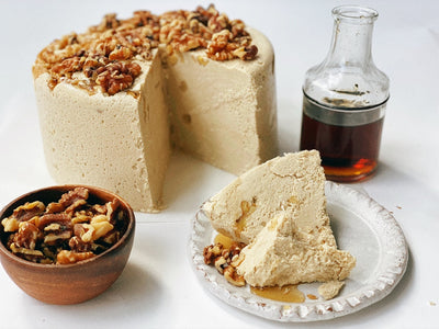 A maple walnut halva cake with a plate of halva, a bowl of walnuts, and a small glass jar of maple syrup